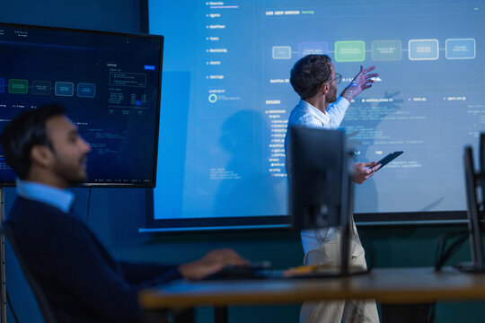A smartly dressed male engineer in a navy blazer at his monitors rests a hand on the mouse while reading through a long log file inside a modern digital office for a global software business
