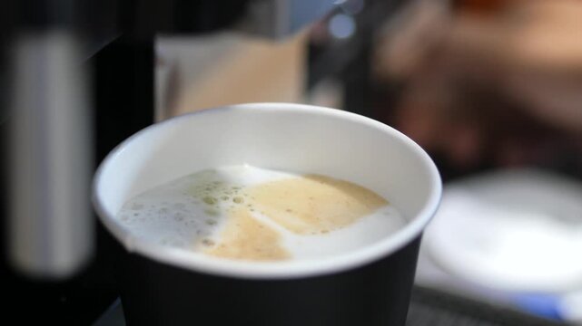 Close up view of freshly brewed coffee with frothed milk in black paper cup with white rim, dynamic beverage making process