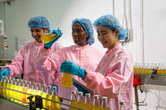 Group of Asian and black factory worker in uniform talking and inspecting rows of glass bottles on the automated conveyor system in manufacturing assembly line.