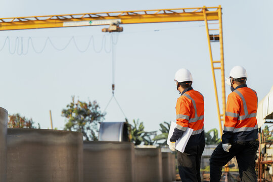 Engineers control crane lifting prefabricated metal cylinder at construction factory yard. Concept of heavy manufacturing, rigging operations, industrial logistics and engineering management.