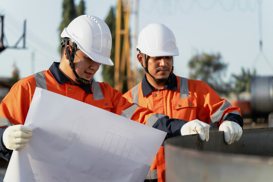 Engineers in hardhats inspect steel pipe with blueprint at fabrication outdoor factory yard. Concept of infrastructure development, quality control and engineering management.