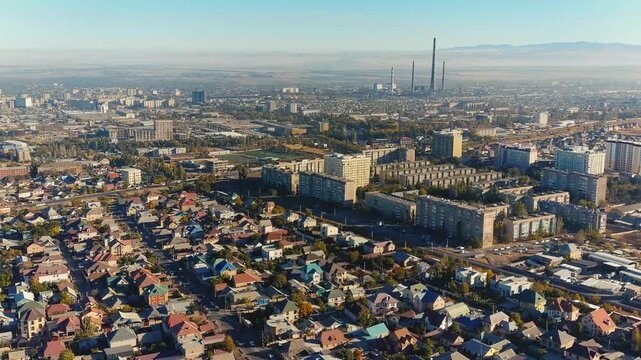 Aerial panorama of Bishkek, Kyrgyzstan, showing residential neighborhoods beside factories and a tall chimney within the urban landscape.
