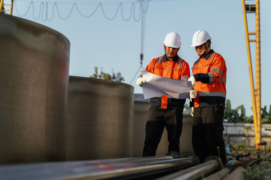 Engineers in hardhats review blueprint under gantry crane at outdoor factory yard. Concept of infrastructure development, civil engineering, and construction planning.
