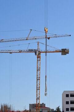 A yellow construction tower crane with long steel cables hanging from its hook alongside another crane jib under clear blue sky