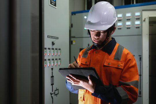 Asian young electrical engineer recording operation metrics on digital tablet screen indoor in control room, Young engineer in white hard hat and orange safety jacket holds tablet and pen