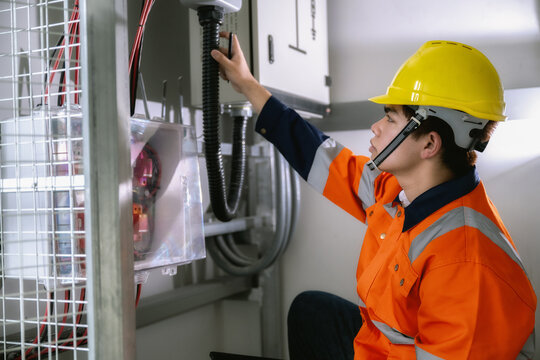 Young technician wears orange safety suit and yellow hard hat. He checks solar inverter cables with a tablet. Focused expression shows careful work. Industrial setting includes metal enclosures