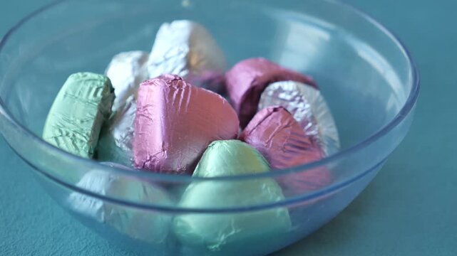 Heart shaped chocolate candies in colorful foil wrappers inside clear glass bowl on blue surface