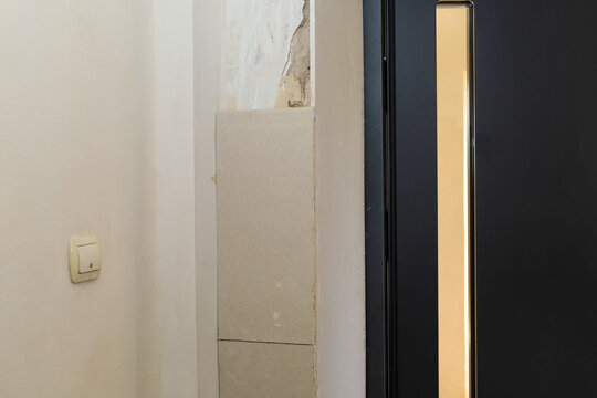 A wall section next to a modern dark door undergoes renovation and drywall installation process. The surface shows unfinished plasterwork, exposed seams, and an installed light switch.