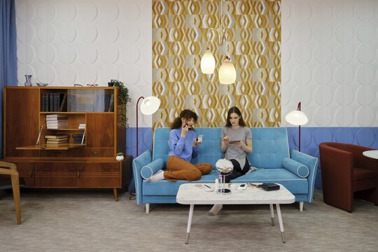 Two friends are seated on a vibrant blue couch in a modern living room, engaged in light conversation while applying make-up