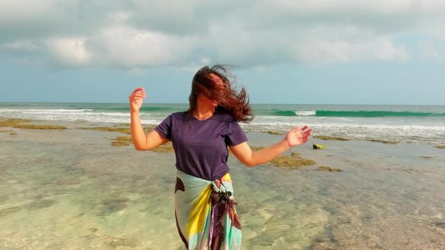 Slow motion medium shot of woman standing in shallow sea water, touching windblown hair on tropical beach with ocean and clouds background.
