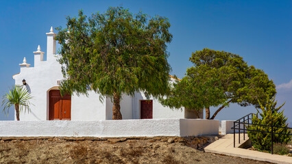 Murcia, Spain - Small whitewashed chapel with simple architecture and a stepped bell gable stands under a bright blue sky in a Mediterranean coastal setting. Chalky white stucco walls are shaded by ta © GertJan