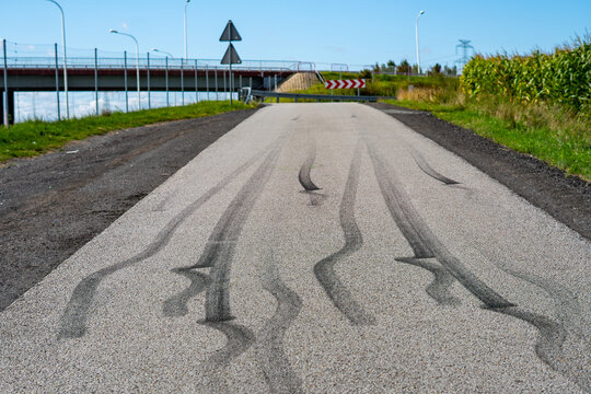 Low-angle shot of black tire skid marks on a narrow asphalt road leading towards a highway overpass bridge, featuring road signs, street lights, safety barriers, and a green cornfield under a clear bl