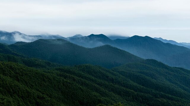 Low clouds drift across layered forested mountain ridges, weaving through green valleys beneath pale overcast sky as distant peaks fade gradually into soft atmospheric haze