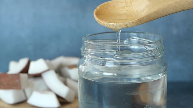 Coconut oil pouring from wooden spoon into glass jar against blue background with fresh coconut pieces