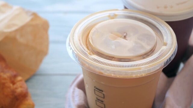 Close-up cinematic shot of two iced coffee drinks with croissant in paper bag on a light blue wooden table, ready for takeaway