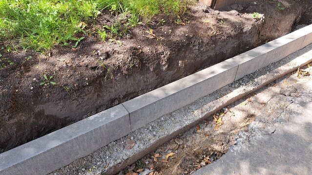 Concrete curb installation along a dirt trench with grass and soil visible, showcasing construction progress on a sidewalk or road project