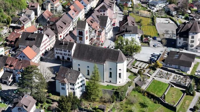 cemetery grounds green lawn headstones border white parish church clock tower spire terraced rooftops old town square sargans streets switzerland aerial overhead drone footage heritage site