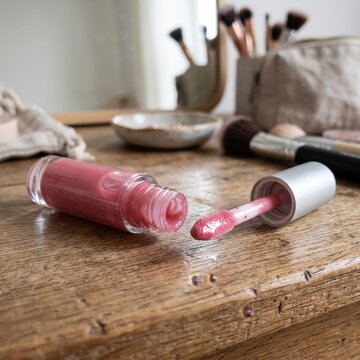A bottle of pink lip gloss lies open on a rustic wooden table, its applicator wand revealing a creamy texture, surrounded by makeup brushes and a cosmetic bag in a softly lit, personal vanity setting.