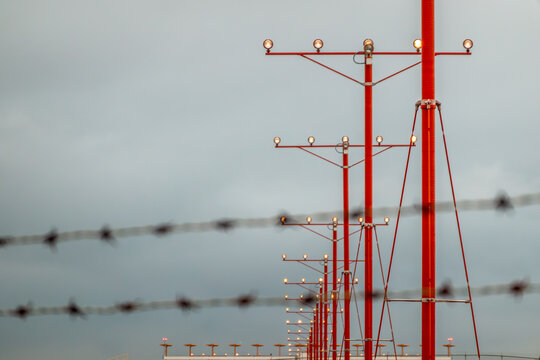 Commercial aircraft on final approach framed by security barbed wire fencing and bright red airport approach lighting systems