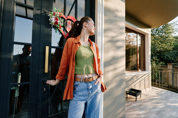 Young woman in orange shirt and green top standing at a black door with a heart wreath. Side view...
