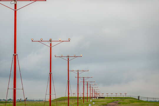 Bright red runway approach lighting towers receding into the distance alongside security barbed wire fencing under an overcast sky