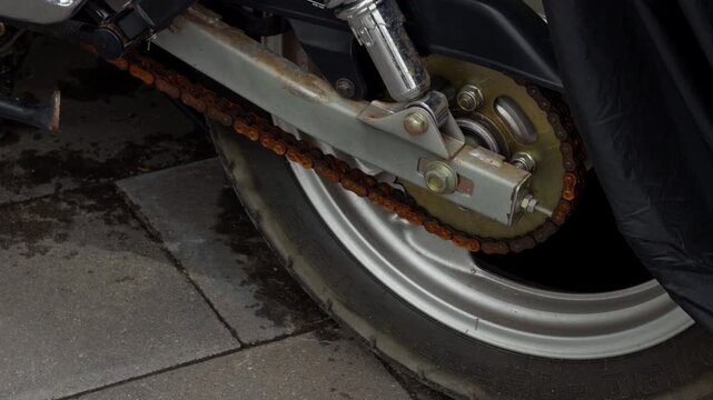 Close-up motorcycle rear wheel with rusty drive chain, sprocket, swingarm, and worn tire on pavement