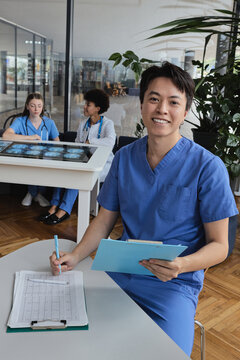 Young Asian doctor in scrubs taking notes over an ECG paper while team members study brain tomography slices