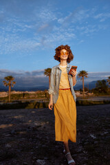 Fototapeta premium woman outdoor lifestyle portrait casual fashion sunny nature backdrop palm trees summer smiling device wind breeze color tone earthy calm leisure confidence denim jacket dress candid moment