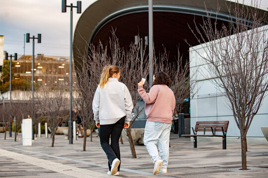 Two young women walking side by side along a paved footpath near a metro station