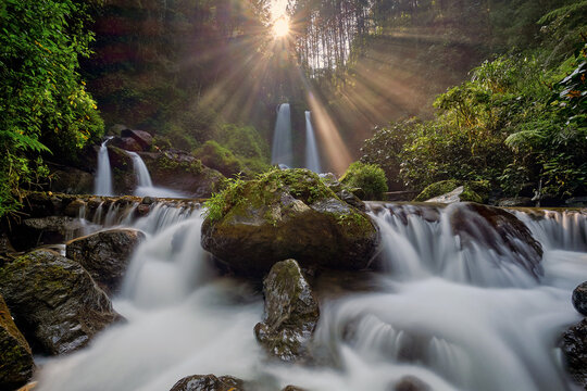 View of Air Terjun Grenjengan Kembar with cascading water, mossy rocks, and sun rays filtering through the forest canopy in Pakis, Central Java, Indonesia.