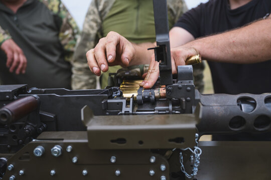 Close up view of a soldier in camouflage uniform using both hands to load a linked ammunition belt into a mounted heavy machine gun.