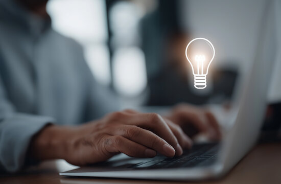Person types on a laptop keyboard at a wooden desk with a glowing lightbulb graphic floating above the device.