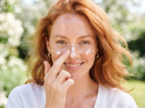Smiling redhead woman applying sunscreen sunblock lotion on nose outdoors