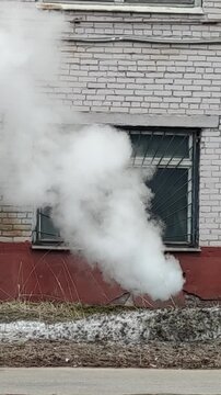 Heavy white smoke and steam escaping from a basement drainage pipe of an old brick public building with windows, depicting a city infrastructure utility failure and emergency accident.