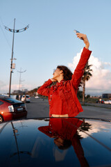 Obraz premium A woman in a bright red jacket takes a dynamic selfie beside a car on a sunny street, with palm trees and a blue sky, conveying urban style and travel energy.
