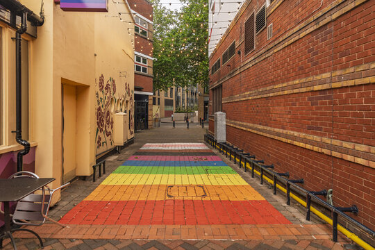 View of a vibrant rainbow-painted brick alleyway with string lights overhead and red brick walls in Birmingham, England, United Kingdom.