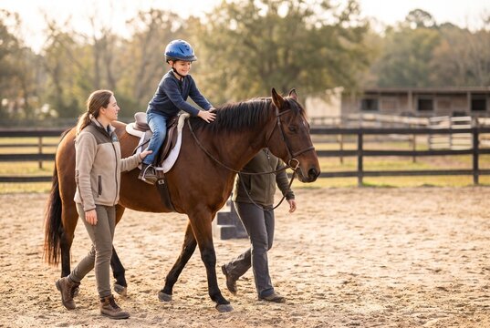 miling little boy in blue helmet riding brown therapy horse led by two hippotherapy instructors in outdoor sandy arena at golden hour sunset, equine assisted therapy and autism rehabilitation concept