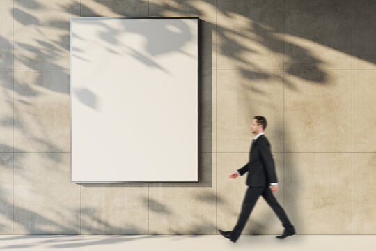Vertical blank poster frame mockup on clean building facade with businessman walking through sunlight and shadow for advertising concept.