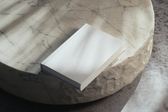 Minimalist 3D render of a blank white hardcover book lying on a round beige marble table with a rough raw edge, soft natural sunlight, botanical window gobo shadow and copy space for book cover mockup