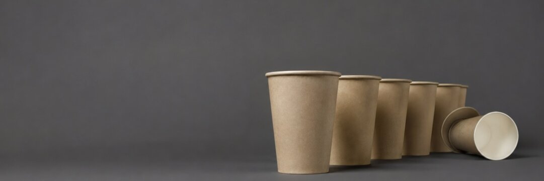 Row of plain kraft paper cups receding across a dark grey studio backdrop with one cup tipped on its side for takeaway drink packaging and branding mockups