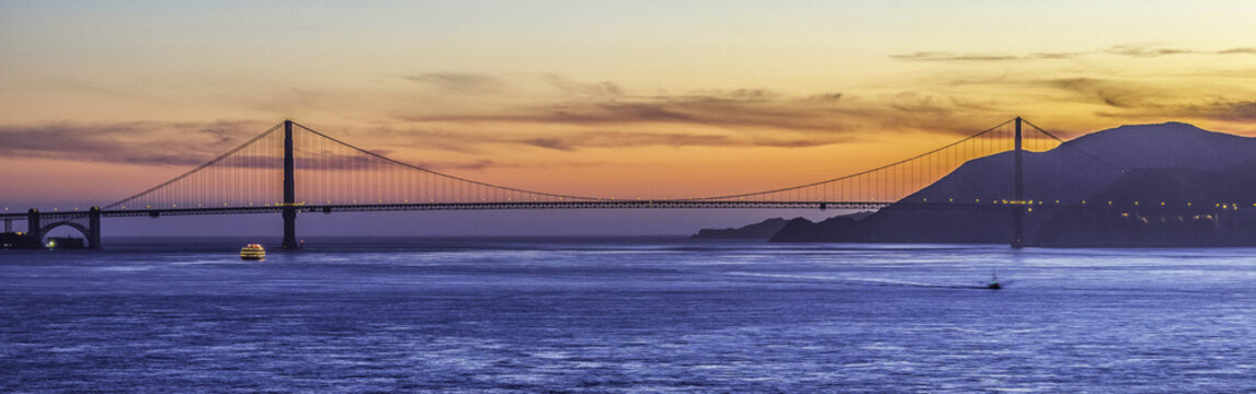 View of the Golden Gate Bridge spanning the bay under a vibrant sunset sky with calm waters and distant hills in San Francisco, CA, United States.