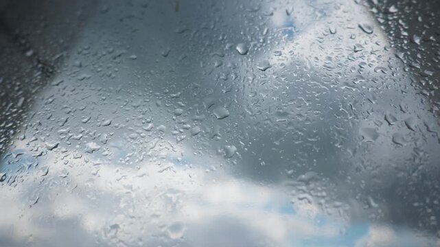 Fresh rainwater beads cling to transparent glass as storm clouds gather overhead. Diffused daylight highlights the organic liquid pattern across the smooth surface.