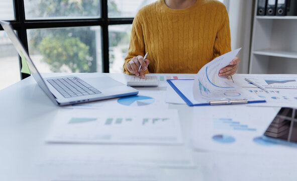 Business professional analyzing financial data and calculating expenses, working on a laptop at an office desk with charts and reports for accounting and auditing