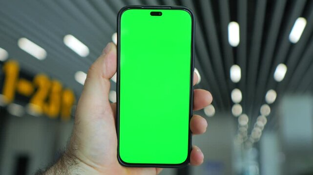 Hand holds phone with green screen. Background shows airport terminal lights. Blurred signage and ceiling lights visible