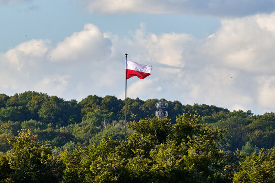 Red and white Polish national flag fluttering on a tall flagpole against clouds and green hills in Gdansk, Poland