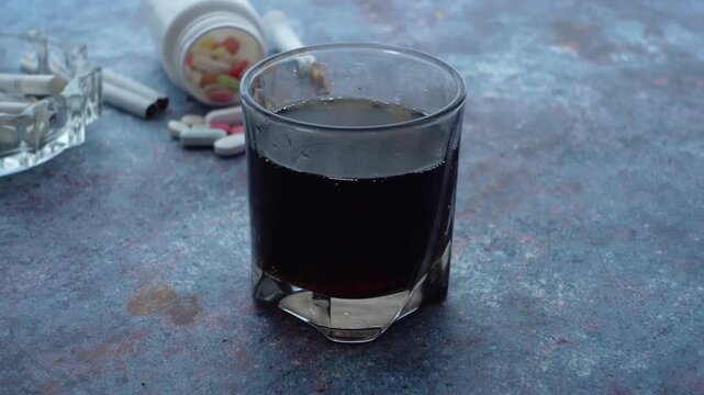Close up of cigarettes, pills, and a glass of dark liquid on a textured surface