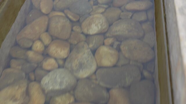 A white flower floats briefly on clear water above smooth pebbles in a wooden garden trough before drifting out of frame.