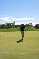 Obraz premium Senior golfer walking confidently across the green with his club in hand, flag and hole behind him - satisfaction and active retirement lifestyle.