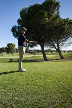 Bearded senior golfer raising his club to aim and line up the direction of his next shot, planning strategy on a tree-lined course.