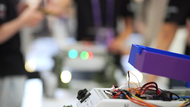 Close-up of electronic device with wires and blue plastic component. Background shows blurred people and colorful lights. Focus on technical details, workshop or tech event atmosphere